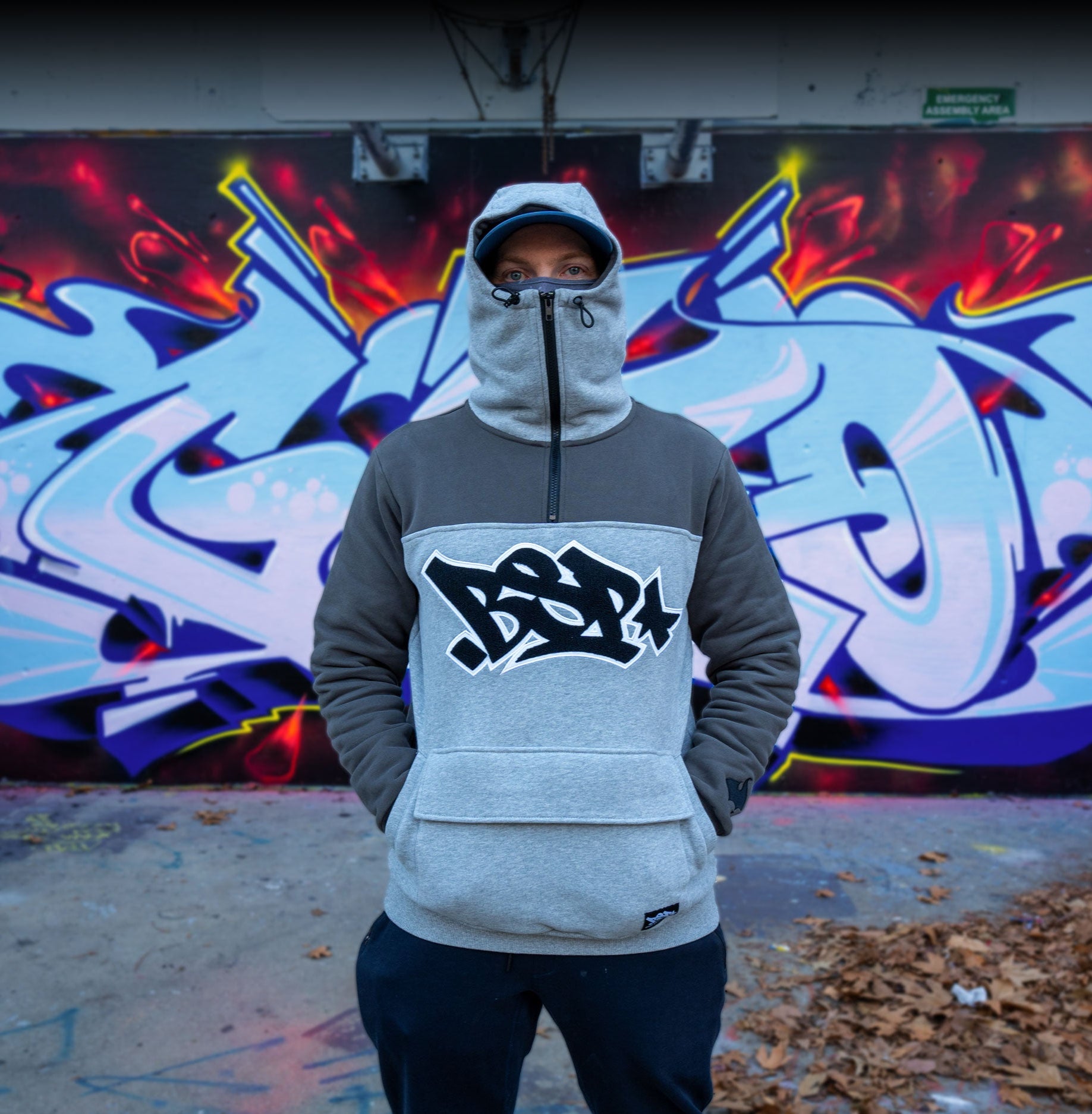 Person wearing a hoodie with a logo in front of a colorful graffiti wall.
